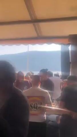 The video shows a crowd of people under a covered outdoor area, possibly a restaurant or bar, with a view of a lake and mountains in the background. The scene is brightly lit by sunlight streaming in from the lake. People are standing around, some are holding drinks. A couple stands near a doorway, embracing. A waiter walks past them. Some people are wearing hats.