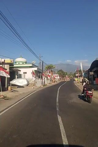 The video pans down a paved road lined with buildings and trees on the left side and a grassy verge on the right. Several red and white flags, possibly national flags, are prominently displayed along the roadside, suggesting a celebration or national event. A mosque with a dome is visible on the left. Power lines are strung across the sky. The road is empty of vehicles and pedestrians. The sky is clear and blue, and mountains are visible in the distance. The scene is lit by bright sunlight.