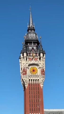 A video shows a tall, ornate tower against a clear blue sky. The tower is made of red brick and features a complex architectural design with multiple levels, spires, and decorative elements. A large, colorful clock face is visible on one of the tower's sections, adorned with red, yellow, and blue patterns. The top of the tower culminates in a slender, pointed spire. The camera angle is from below, looking up at the tower. The video is extremely short, lasting only a few seconds, and appears to be a static shot with no movement.