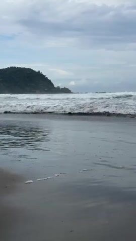 The video shows a slow pan of a beach. The foreground is dominated by wet sand and shallow water reflecting the cloudy sky. In the midground, waves are breaking and rolling towards the shore. The background features a lush green hill with some buildings visible on its slope. The sky is overcast with grey clouds. There are no people or distinct objects other than natural elements.