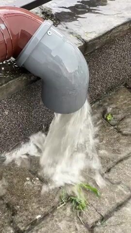 Water is gushing out of a gray downspout which is attached to a larger brown pipe. The water is splashing onto a paved surface made of interlocking gray stones. Some green blades of grass are visible on the ground, with some of them being pushed by the flowing water. The scene appears to be outdoors, likely during or after rain.