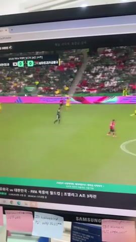 A screen displays a soccer match. The score is 0-0. Players in red and yellow uniforms are on the field. Spectators fill the stands. The screen also shows a search bar and a live broadcast information banner. Below the screen are sticky notes and a part of a Samsung monitor.