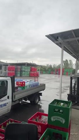 The video displays an outdoor scene at what appears to be a beverage distribution center or warehouse yard in Kovachevo, Bulgaria. The weather is overcast and rainy, with raindrops visible and the ground wet.

In the foreground, several green and red plastic crates, many containing empty glass bottles, are stacked. A prominent green crate displays the text "ПИРИНСКО," indicating a Bulgarian beer brand.

To the left, a white pickup truck, possibly a Ford Transit, is parked. Its flatbed is loaded with various crates of bottled beverages. Red crates with "Coca-Cola" branding are visible, as are bottles of "Банкя" (Bankya), a Bulgarian mineral water. Text on the side of the truck reads "АРД ООД" and a partial phone number "00 35 21 22."

In the middle and background, numerous large stacks of red and green crates are arranged across the wet yard, suggesting a substantial inventory of bottles. Some of these stacks are under a large metal canopy or shelter on the right side of the frame, where a forklift is also partially visible. The overall setting is industrial, with no people actively engaged in work within the shot, possibly due to the rain.