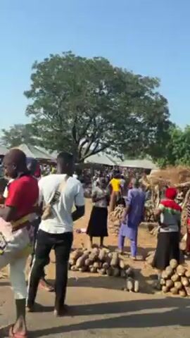 The video shows a scene captured from a moving vehicle, possibly in a rural area or small village. On the side of the road, there are various wooden structures, some appearing to be stalls or shelters with thatched roofs. People are visible around these structures, some standing and seemingly engaged in trade or conversation. A motorcycle is parked on the side of the road and there are some other cars driving by as well. The environment appears dry and dusty, with trees dotting the landscape under a clear, blue sky.
