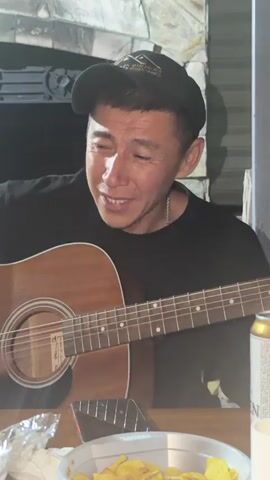 A man wearing a black t-shirt and a black baseball cap is playing an acoustic guitar. He is looking down at the guitar with his eyes closed and a smile on his face. The background appears to be an outdoor setting with a stone wall and some dark foliage. There are also some objects on the table in front of him, including a can, some chips, and a cell phone. The lighting is dim, suggesting it is nighttime.