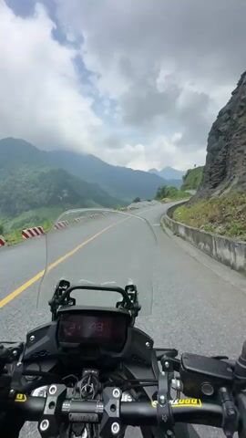 The video provides a first-person perspective of a motorcycle ride through a scenic, mountainous landscape in Minh Sơn Commune, Vietnam. The camera is mounted on the motorcycle, showing the top of the windshield, handlebars, and a digital dashboard displaying "HONDA" and "31:48," which likely indicates time or speed.

The motorcycle is traveling on a paved asphalt road, characterized by a double solid yellow line in the center and a white solid line on the shoulder. Red and white striped barriers are visible along the left side of the road, behind which loose stones are scattered. On the right, a steep, rocky mountain face rises immediately beside the road.

The surrounding environment is dominated by lush green mountains, some of which are partially shrouded in clouds, suggesting a high elevation and possibly misty conditions. The sky is largely overcast, but the lighting indicates daytime. The road curves gently to the right, inviting exploration of the expansive natural scenery. The continuous sound of the motorcycle engine confirms steady movement along this picturesque mountain pass.