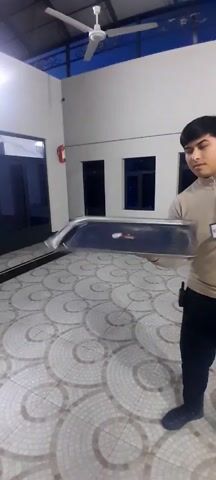 A young man wearing a light brown long-sleeved shirt and black pants stands indoors. He holds a large, rectangular, silver metal tray, twirling it at first and then holding it stationary in front of him. The room has a tiled floor with a circular pattern, white walls, and several windows, some covered and some revealing a dark exterior. A fire extinguisher is mounted on the wall. The man wears a name tag and what appears to be a small radio device on his belt.