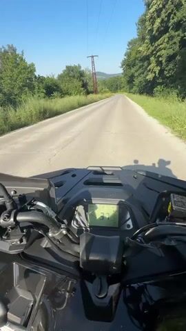 This media file captures a first-person perspective of someone riding an ATV on a paved road in Orlovtsi, Bulgaria. The scene is set on a sunny day, with a clear blue sky. The road is flanked by lush green grass and trees, indicating a rural or semi-rural setting. In the distance, power lines and a red electrical tower are visible, suggesting the presence of infrastructure in the area.

The rider's point of view shows the ATV's handlebars and digital dashboard, which displays the speed as "50". A shadow of the rider and ATV is cast on the road ahead. In a brief cutaway, a person wearing sunglasses and a white t-shirt is seen looking towards the camera, giving a glimpse of the rider. The sun is bright, creating lens flare in some shots. The overall mood appears to be one of enjoyment and leisure, characteristic of an outdoor recreational activity. The time of day is clearly daytime due to the bright sunlight and blue sky.