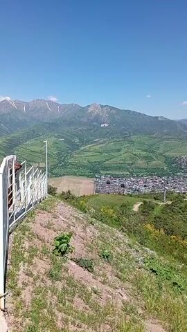 The video shows a wide, panoramic view of a town nestled in a valley surrounded by green hills and mountains. The camera is positioned on a hillside, looking down at the town below. In the foreground, there is a white fence and some sparse vegetation, including grass, small plants, and some yellow flowers. Beyond the foreground, the view opens up to reveal the town, which appears densely built with low-rise buildings. The town is situated in a valley with patches of greenery and what appears to be a lake or river visible on the right side. In the distance, snow-capped mountains rise against a clear blue sky. The video appears to be taken on a sunny day, providing good visibility of the landscape.