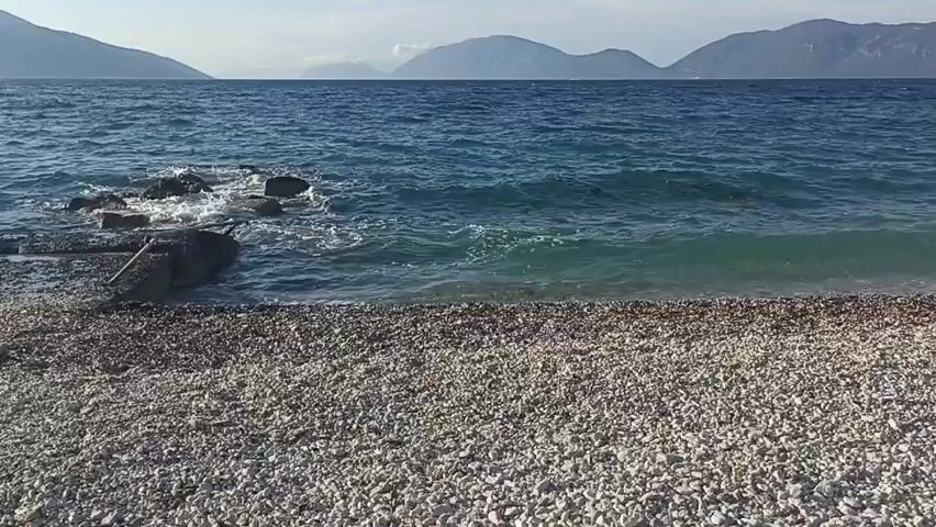 A wide shot shows a pebble beach in the foreground. The ocean is to the left and center, with gentle waves lapping at the shore. In the distance, mountains rise above the water under a pale sky. A small rocky outcropping with some debris is visible in the water near the left side of the frame. The scene is calm and appears to be a natural coastal environment.