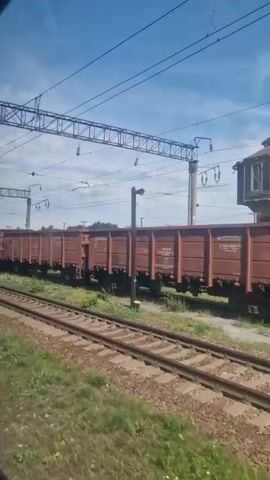 The video shows a train traveling alongside a long line of freight cars. The freight cars are dark red and appear to be open-top gondola cars. The train is moving along a double track, with grassy areas and trees on either side. In the background, there is a tall, wooden water tower and power lines are visible overhead. The sky is blue with some clouds. The perspective is from inside another train, looking out a window.