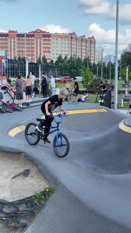 A group of people, mostly young males, are gathered at a skate park. The skate park features a pump track with sculpted asphalt mounds and yellow racing stripes. Several individuals are on bicycles and scooters, some actively riding on the track, while others are standing or sitting nearby. In the background, there are modern apartment buildings, trees, and a cloudy sky. The overall atmosphere appears to be one of recreation and outdoor activity.