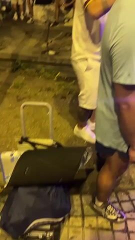The video shows a group of people gathered outdoors at night, possibly in a public square or street.  There is a large stone monument in the background.  Several people are visible in the foreground and midground, some of whom appear to be dancing or interacting.  One person in the foreground is wearing a light blue t-shirt and a cap, holding a drink. To their left, a person in a white t-shirt and white shorts is seen from behind.  Further back, several other individuals are seated or standing, some illuminated by lights. The overall atmosphere appears to be festive.