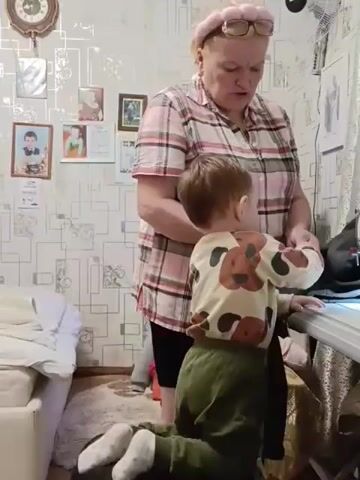 The video shows an elderly woman in a pink and white plaid shirt standing next to a toddler in a brown and beige animal print sweatshirt and green pants. The toddler is kneeling on the floor near the woman and a white sewing machine. The woman appears to be instructing or showing the child something on or near the sewing machine. The room is cluttered with photos, a clock, a fan, and other items.