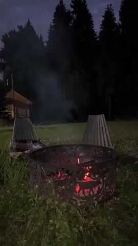 A campfire crackles in a metal fire pit at dusk, with smoke rising into the dark sky. In the background, a small wooden structure and two conical wooden objects are visible. The scene is set in a grassy area surrounded by trees. The fire pit has a decorative cutout pattern featuring deer.