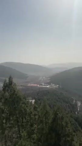 The video shows a hilly landscape with a city in the distance. The sky is a pale blue and the sun is shining. There are trees in the foreground and the hills are covered in green vegetation. The city in the distance is hazy. There are people talking in the background, although they are not visible. The camera pans slowly from left to right.
