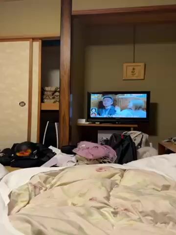 The video shows a bedroom with a rumpled bed in the foreground. On the bed are various items including a black backpack, a pink scarf, and clothing. A television is positioned on a shelf in the background. The TV screen displays a Japanese TV show with two people being interviewed. To the left of the screen is a storage area behind a sliding door. The room has neutral colored walls and a framed piece of artwork hangs above the TV.