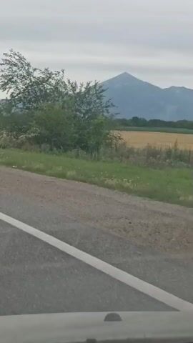The video captures a scenic view from a moving vehicle. The foreground shows a paved road with white lane markings. To the right of the road, there is a field of yellow crops and green vegetation. In the background, a large, imposing mountain range dominates the horizon under a cloudy sky. The camera also briefly pans to show a green guardrail alongside the road and a distant transmission tower on top of a hill. At one point, a dark-colored car is parked on the shoulder of the road with two people standing nearby.