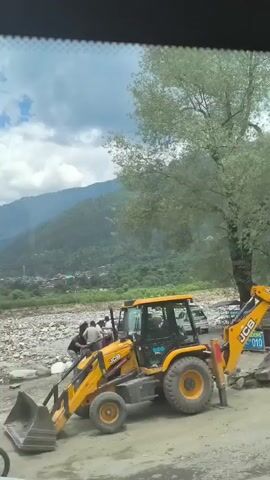 The video captures a scene on a road next to a rocky riverbed with a mountain range in the background. In the foreground, a yellow JCB excavator is parked, with several people gathered around it. Some individuals appear to be working on or near the excavator, while others are standing and observing. A silver car is partially visible in the lower right corner. The environment is rural and appears to be undergoing some form of construction or road maintenance. The sky is partly cloudy with a clear blue sky visible in sections. There are also yellow and black traffic barriers present. The overall impression is one of activity related to heavy machinery in a mountainous, natural setting.