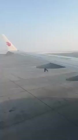 The video shows a plane wing with a red and white logo on its winglet, possibly a China Southern Airlines plane, on a gray concrete tarmac. The weather is hazy, with a light blue sky. The camera is positioned inside the plane, looking out. The plane is stationary.