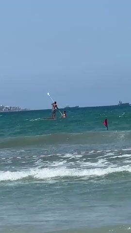 Two people are visible in the ocean. One person, appearing to be male, is standing on a paddleboard and holding a paddle. Another person, whose gender is not clear, is in the water behind the paddleboarder. A red flag is planted in the ocean. In the background, there are several large ships and a distant coastline. The water is green and has waves. There is a red and white rope floating in the foreground, likely marking a boundary. The sky is clear and blue.