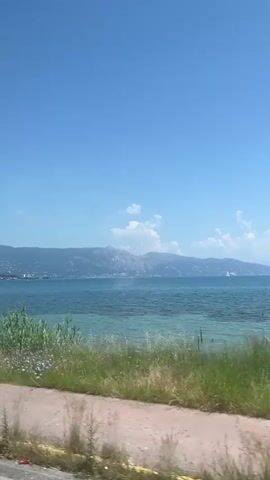 The video shows a view of a coastline with a body of water and mountains in the background, taken from inside a moving vehicle. The sky is a clear blue with scattered clouds. The water transitions from a light turquoise near the shore to a deeper blue further out. There is a small sailboat visible in the distance. A strip of land is visible alongside the water, covered in vegetation. The road surface is partially visible, with a section of a car's roof and back window in the lower-left corner. The video appears to be taken during daylight, with sunny conditions.