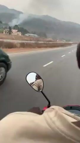 The video appears to have been recorded from a motorcycle driving on a highway. The camera is focused on the road ahead and the motorcycle's side mirror. In the distance, there's a green minivan driving in the same direction.  Beyond the road, there is a hillside with patches of smoke, suggesting a fire. The person riding the motorcycle points towards the smoke. There are a few other vehicles on the road. The sky is overcast, and the overall environment is somewhat hazy.