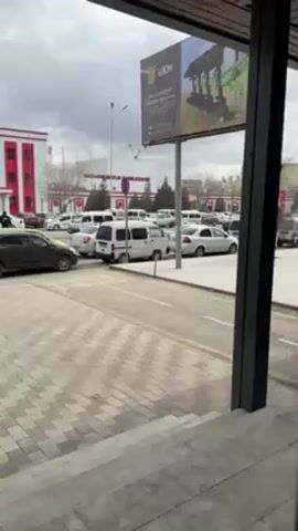 The video shows a street scene outside a building with a Coca-Cola sign. There is a parking lot full of cars, a billboard, and a sidewalk made of gray brick-like pavers. The sky is overcast. A person in a dark coat walks amongst the parked cars. The video is shot from an elevated position, possibly from inside a building looking out a window or doorway.
