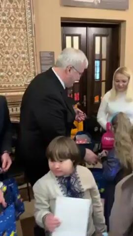 The video shows a man in a black suit handing out gifts from a blue bag to children and their families. The children appear to be dressed up for a special occasion. The background includes an ornate tiled wall, a wooden door, and informational panels. One child is wearing a white shirt with Ukrainian embroidery. Another child wears a Santa hat. The environment appears to be inside a building with a patterned tile floor. The interaction seems to be festive and welcoming.