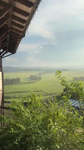 A sweeping, panning shot reveals a rural landscape at dawn or dusk. The foreground is dominated by lush green foliage and the edge of a wooden structure with a gabled roof, suggesting an outdoor seating area or balcony. In the mid-ground, rolling green fields stretch out, dotted with trees and patches of mist that add a soft, atmospheric quality. A low-lying building, possibly a house or barn, with a dark roof and wooden siding, is visible on the right side of the frame, partially obscured by branches. The sky above is a mix of soft blues and whites, with the sun casting a warm, bright glow through the clouds, indicating either sunrise or sunset. The overall impression is one of tranquility and natural beauty.