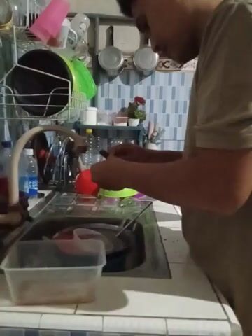 A person is washing dishes in a kitchen sink. The individual is wearing a tan shirt and has dark hair. They are focused on cleaning a red cup under the running water from a faucet. The sink is located next to a dish rack filled with various kitchen items, including cups, bowls, and plates. Behind the sink, there is a blue and white tiled wall. On the wall, there are three metal pots hanging, and a small shelf with a potted plant and other items. There is a bottle of water on the counter.