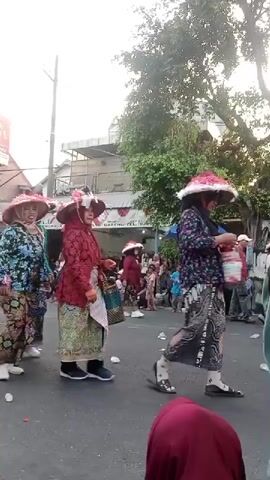 A street parade is underway. Several people are walking in procession, adorned in traditional clothing and wide-brimmed hats. The individuals in the foreground appear to be women wearing vibrant, patterned garments, including skirts and blouses. They are walking on an asphalt road. On the left side of the road, there are spectators gathered, and some structures, including a building with a banner and a billboard. On the right side, there are more spectators, trees, and what appears to be a blue umbrella. The sky is visible in the background, suggesting it is daytime. The overall atmosphere seems festive.