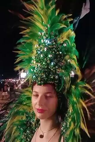 A woman in a green feather headdress and a green sequined top stands in front of a busy background at night. She is facing forward, looking slightly to the left, and smiling. She has a short haircut and is wearing a necklace. Her makeup is visible, with pink lipstick and some glitter on her cheekbones. In the background, other people are visible, some walking, and there are what appear to be tents or stands with lights. The scene is outdoors and appears to be a night market or a festival.