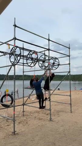 Two men are participating in an obstacle course on a sandy beach next to a body of water. They are both hanging from large black suspended tires on a metal scaffolding structure. The man on the left wears a blue long-sleeved shirt and dark pants, holding a tire with both hands while his feet rest on a lower bar. The man on the right wears a black t-shirt and dark shorts, also holding a tire with both hands with his feet on a lower bar. Other people are visible on the beach and in the water in the background under a cloudy sky.