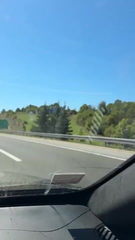 The video is a first-person view from inside a car driving on a highway. The road is paved with asphalt and has white lane markings. On the right side of the road, there is a guardrail. Beyond the guardrail, the landscape consists of a grassy hill with scattered trees. A large billboard is visible on the hill. Further down the road, there is another green road sign with white text and numbers, indicating distances to various locations. The sky is clear and blue. The car's dashboard is visible in the foreground. The video appears to have been recorded on a sunny day.