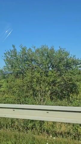 The video shows a view from a moving vehicle. A guardrail is visible in the foreground. Beyond the guardrail, there are green trees and vegetation. The sky is blue with some white streaks, possibly contrails. In the distance, there are rolling hills and scattered buildings, suggesting a rural or semi-rural landscape. The lighting indicates it is daytime.