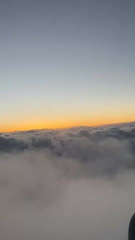 The video shows a view from an airplane window looking out over a dense layer of clouds. Above the clouds is a clear sky transitioning from gray to a thin orange line near the horizon, indicative of either sunrise or sunset. The bottom right corner of the frame shows a portion of the airplane's wing or engine. No people are visible. The overall scene is serene, emphasizing the natural beauty of the sky and cloudscape.