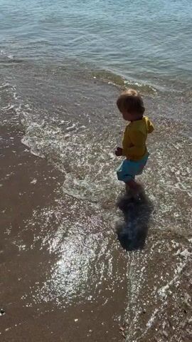 A young child with blonde hair, wearing a yellow long-sleeved shirt and blue shorts, stands near the edge of the water on a sandy beach. The child is facing away from the camera and is partially submerged in shallow water that laps at their feet. Sunlight glints off the water's surface. A dark shadow of the child is cast on the wet sand. The ocean extends into the background under a bright sky.
