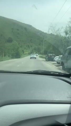 A video captures a view from inside a car driving along a paved road. The road is bordered by a green, mountainous landscape with trees. Several cars are visible on the road, either ahead or in the side-view mirror. A dark-colored car is parked on the right side of the road, and a white car is driving ahead. The sky is overcast. The video transitions slightly, showing the car's interior and the side-view mirror reflecting the passing scenery. The overall impression is of a scenic drive through a hilly, natural environment.