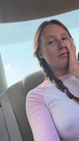 The video shows a young woman with braided hair looking out a car window. The camera then pans to the interior of the car, showing the dashboard, steering wheel, and a rearview mirror with a blue beaded ornament hanging from it. The car appears to be driving on a mountain road, with green hills and blue skies visible through the windows. A seatbelt is also visible.