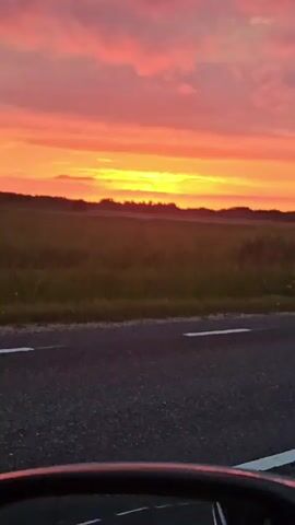 The video shows a scenic view of a sunset over a field, seen from the perspective of a vehicle traveling on a road. The sky is ablaze with vibrant colors of orange, yellow, and pink, with clouds scattered across the horizon. The sun is setting behind a distant treeline, casting a warm glow over the landscape. The foreground of the video features an asphalt road with white lane markings. On the right side of the road, there are some trees and vegetation. The camera appears to be mounted inside or on a moving vehicle, as indicated by the slight motion blur and the perspective.