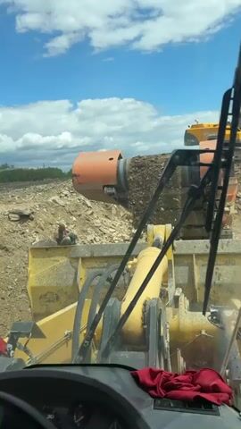 From within a yellow wheel loader, the video shows a construction site. A worker in a green shirt and blue pants is on the ground in front of the loader, appearing to search through a pile of rocks and dirt. Behind him is an orange rotating separator sorting material. An excavator is visible in the background. The weather is sunny, with scattered clouds.