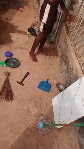The video shows a top-down view of a dirt surface outdoors. Present are a blue dustpan, a small metal bowl, a white rectangular surface resembling a table or countertop with a marbled pattern, and a mop with a blue head and a green-and-white polka-dotted handle. The objects are lying on the ground. The surface is mostly dirt with some concrete bordering it.
