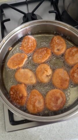 The video shows a stainless steel pan containing about a dozen round, golden-brown fritters frying in oil. The pan sits on a gas stovetop with a black burner grate. Bubbles surround the fritters as they cook. A stainless steel pot is visible in the background to the upper right of the pan. The countertop under the stovetop is a light gray color with subtle patterns.