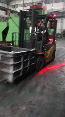 A yellow forklift with a driver is moving through a factory. The forklift has red warning lights on its rear. The scene is indoors, with concrete floors and industrial machinery. There are large green and red machines with yellow safety cages in the background. The lighting suggests it is daytime, as there is artificial lighting present, but no indication of natural light or weather conditions. The overall atmosphere appears to be one of industrial activity. There is some text visible on the red part of the large machine in the background, which appears to read "2000TON."