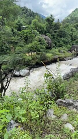 A fast-moving, muddy river flows through a lush, green mountainous landscape under a cloudy sky. The riverbanks are lined with large rocks and dense vegetation, including trees and bushes. In the background, a small building or structure can be seen nestled among the trees on the hillside. The overall impression is one of a wild and natural environment.