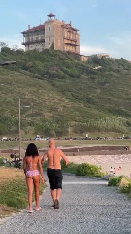 Two people, a man and a woman, walk away from the camera on a paved path. The woman is wearing a purple bikini, and the man is wearing black shorts and is shirtless, revealing a tattoo on his back. They are walking towards a beach with several other people scattered on the sand. Some people are sitting on blankets, while others are standing or playing. There is a green umbrella on the beach. Behind the beach, there is a low concrete structure. Further back, a road runs along the coast with cars parked on the side. Beyond the road, a steep, grassy hill rises, with a large, multi-story building situated near its summit. The sky is partly cloudy with a soft, evening light.
