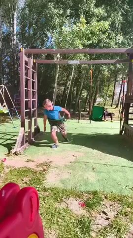 The video shows a boy on a playground. He is wearing a blue t-shirt and patterned shorts. The boy is running towards a swing set, holding a green object, possibly a leaf blower or some kind of tool. He then jumps into the air as he approaches the swing set. In the background, another person is visible, lying on the ground near a slide. The playground appears to be outdoors, with trees and foliage surrounding the area. There is also a red object in the foreground, partially visible.