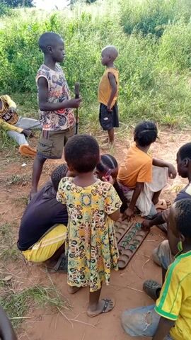 A group of about ten children is playing a game outdoors, in a field of dirt surrounded by green foliage. Several of the children are kneeling or sitting around a game board, which has round depressions in it and contains small, round, green marbles. The children are taking turns moving the marbles around the board. Several children stand around watching the game. One child holds a stick and is talking. The children are wearing casual clothes. The day is sunny.
