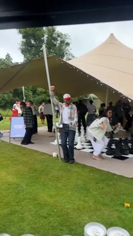 A man with a beard wearing a white tank top, an open plaid shirt, dark pants, and a red and white baseball cap holds a white cup in his right hand, with his left fist raised. He stands next to a white pole, in front of a giant chessboard. He is smiling and looking to his right. In the background, under a large beige tent, several people are gathered around the chessboard, and others are milling about in the grassy area outside the tent. A woman in white pants and a white jacket is visible on the right side of the frame, crouching near the chessboard. The event appears to be outdoors, with trees visible in the background.